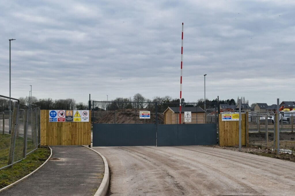 Construction site entrance with closed gate and barriers beside a public road.