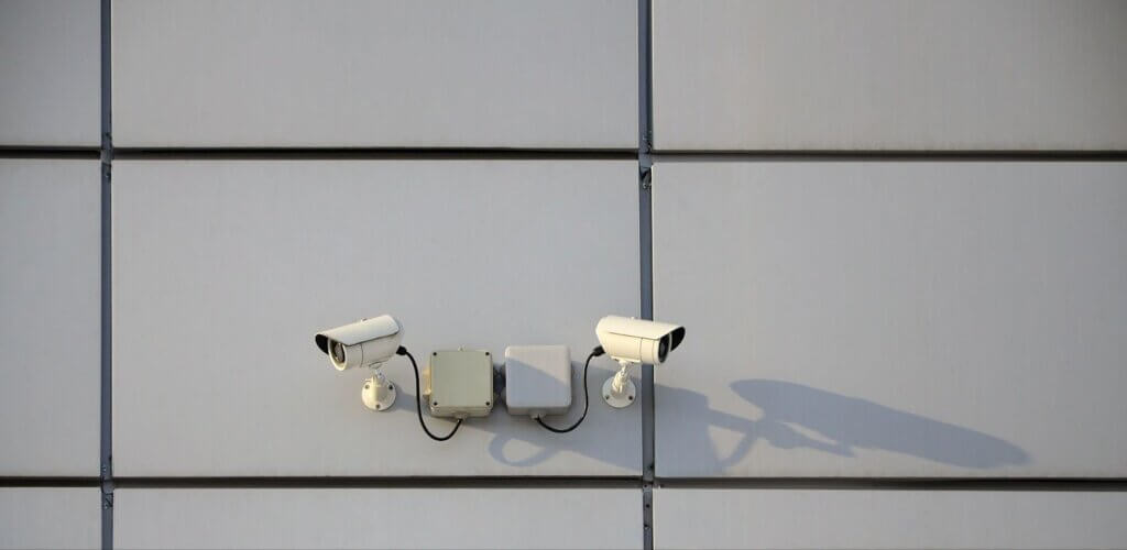 Two white security cameras mounted on a gray metal wall, casting long shadows in sunlight.