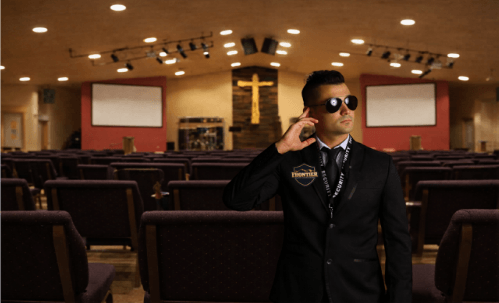 A security guard wearing a suit, sunglasses, and an earpiece stands inside a church sanctuary with rows of seats and a cross at the front.