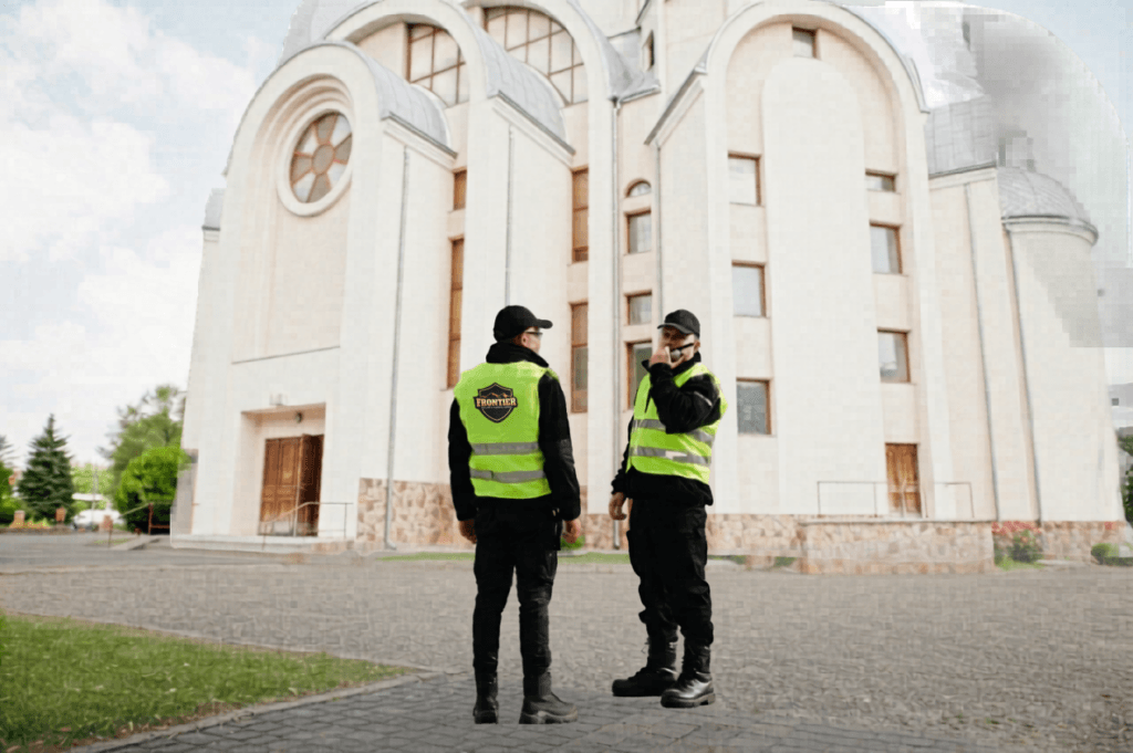 Two security guards in high-visibility vests stand and talk outside a large church building near the main entrance.