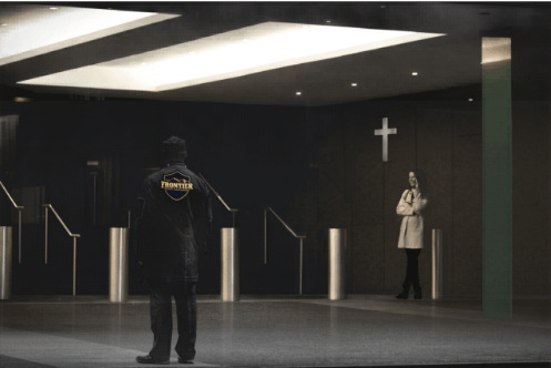 A security guard stands in a dim church lobby facing the entrance area while a woman waits near a wall with a lit cross.