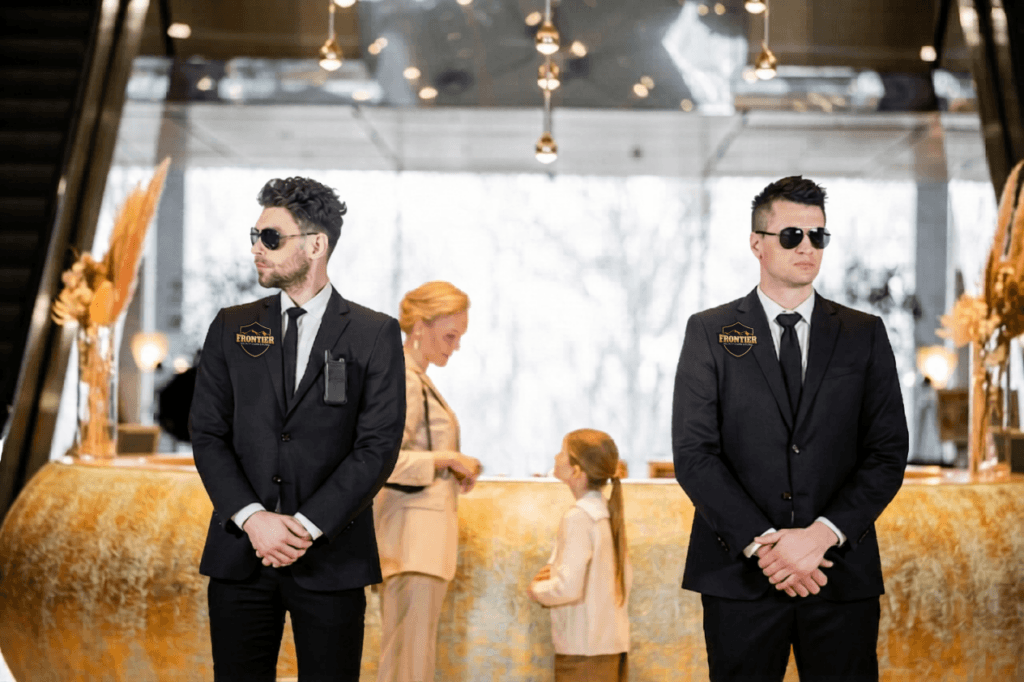 Two hotel security guards in suits stand at the lobby entrance while guests check in at the front desk behind them.