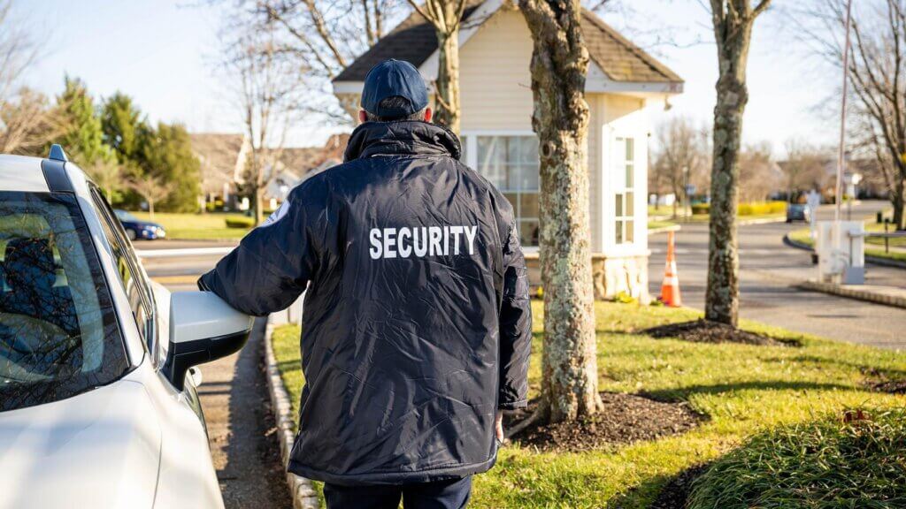 Security guard in a dark jacket labeled SECURITY stands beside a white SUV on a suburban street (rear view).